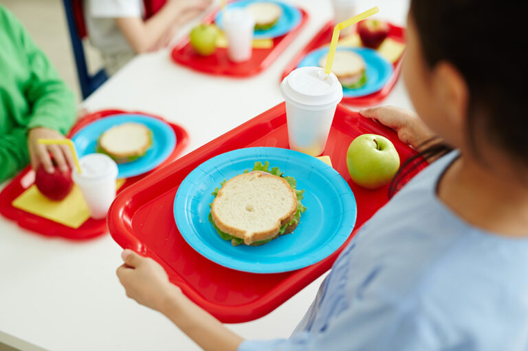 Schoolgirl carrying red plastic tray with sandwich on plate, glass of drink and green apple during lunch break